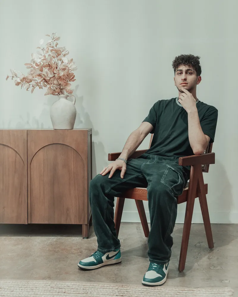 Man in a dark t-shirt and green sneakers seated on a wooden chair beside a sideboard with dried flowers