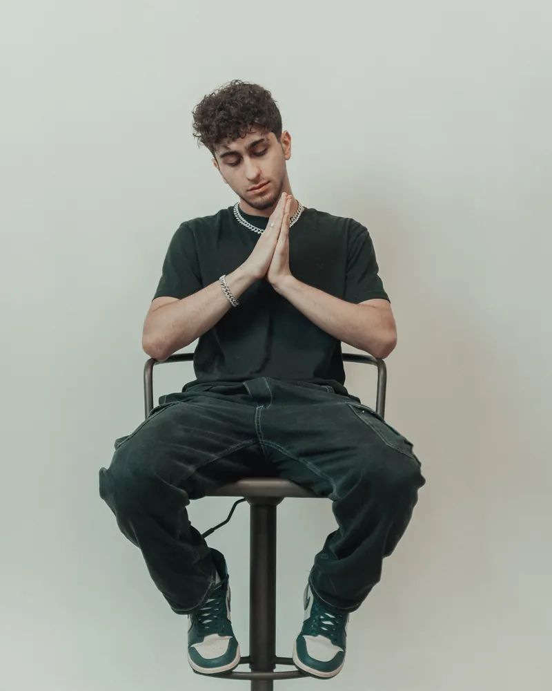 Man in a dark t-shirt seated on a stool with his hands pressed together in front of his chin, against a pale wall