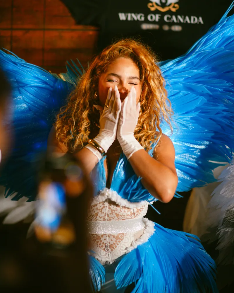 Model in blue feathered wings and white gloves laughing with her hands over her mouth